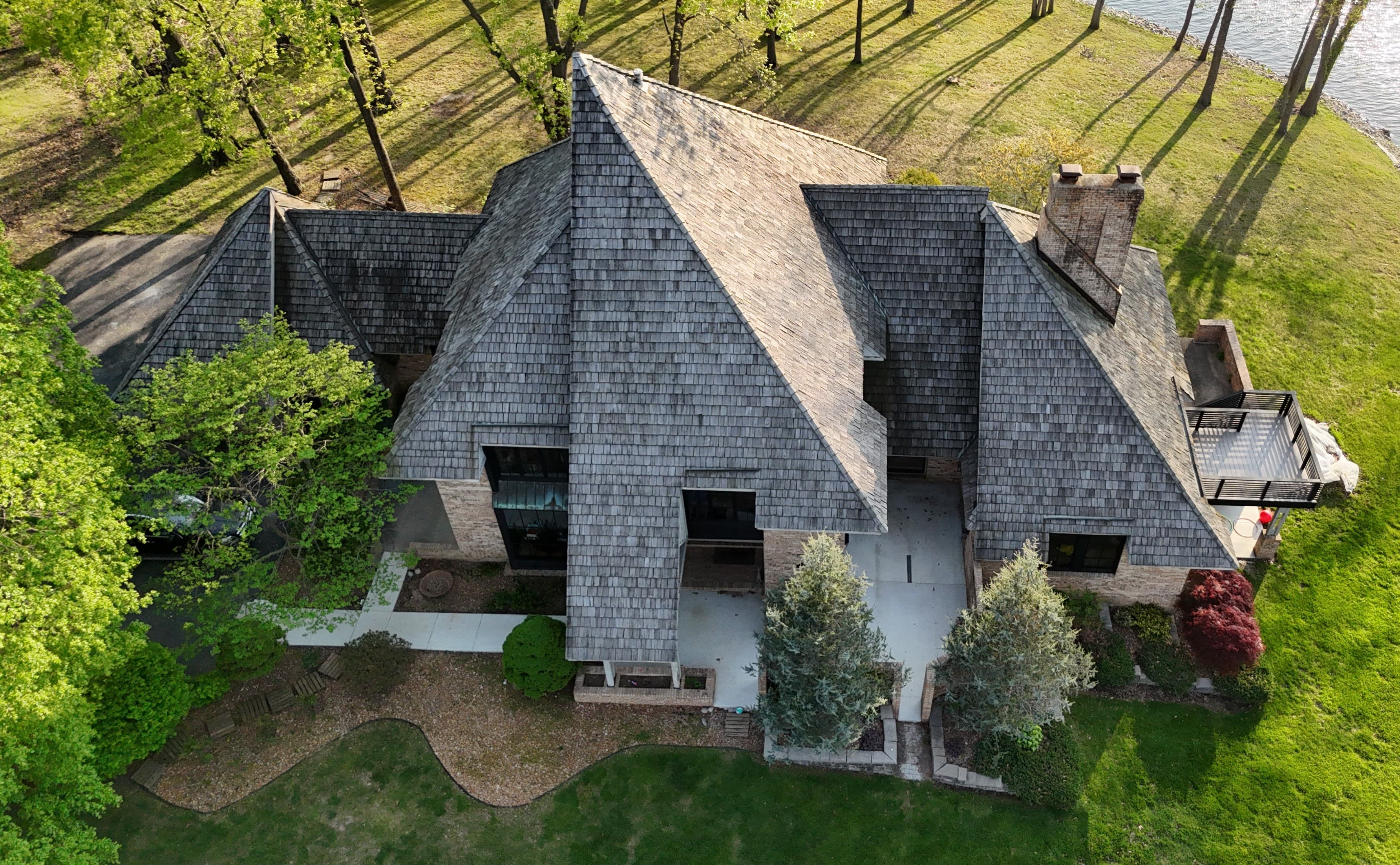 The Architect's Retreat — aerial front elevation before restoration: weathered, faded cedar shake roof on a multi-gable brick residence beside the lake in summer